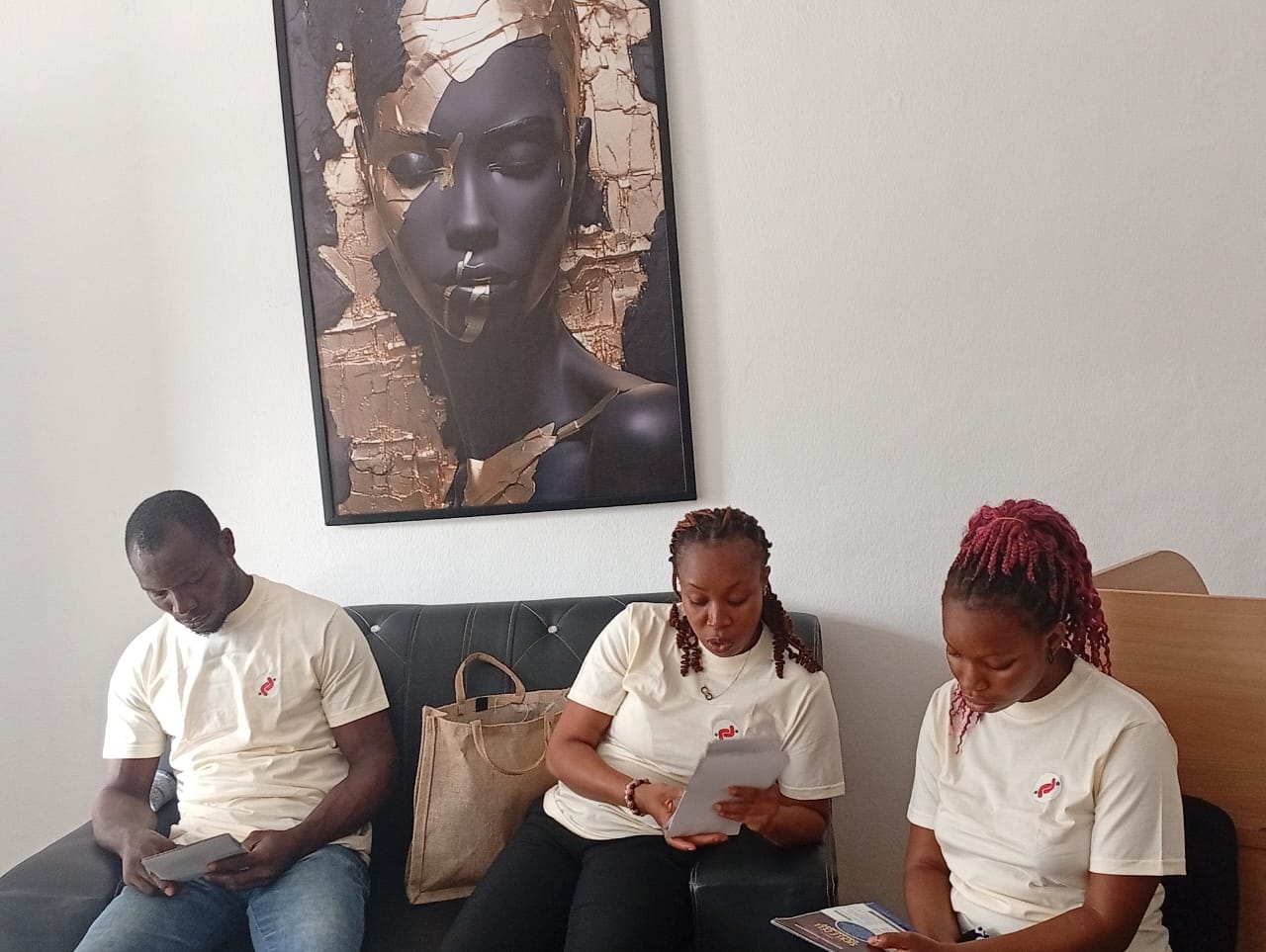 Three people in matching shirts seated on a couch, reading pamphlets, with a large portrait behind them.