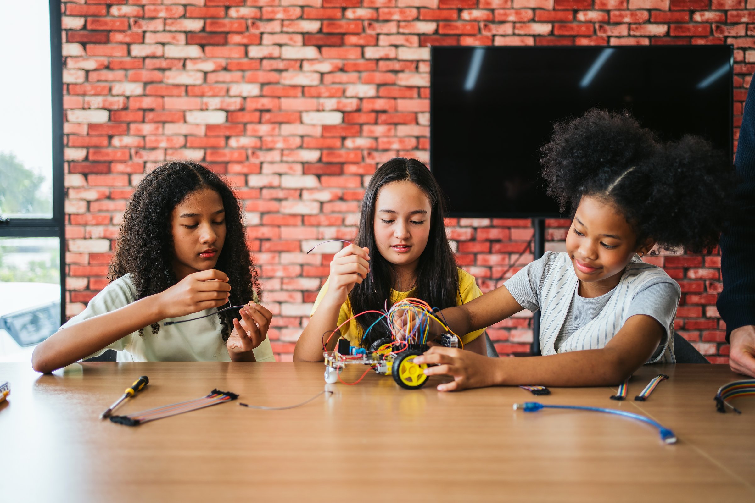 Three diverse schoolgirls building a robotic car with wires in a classroom. Engaging in STEM learning, teamwork, and coding activities. Hands-on education encourages creativity and innovation.