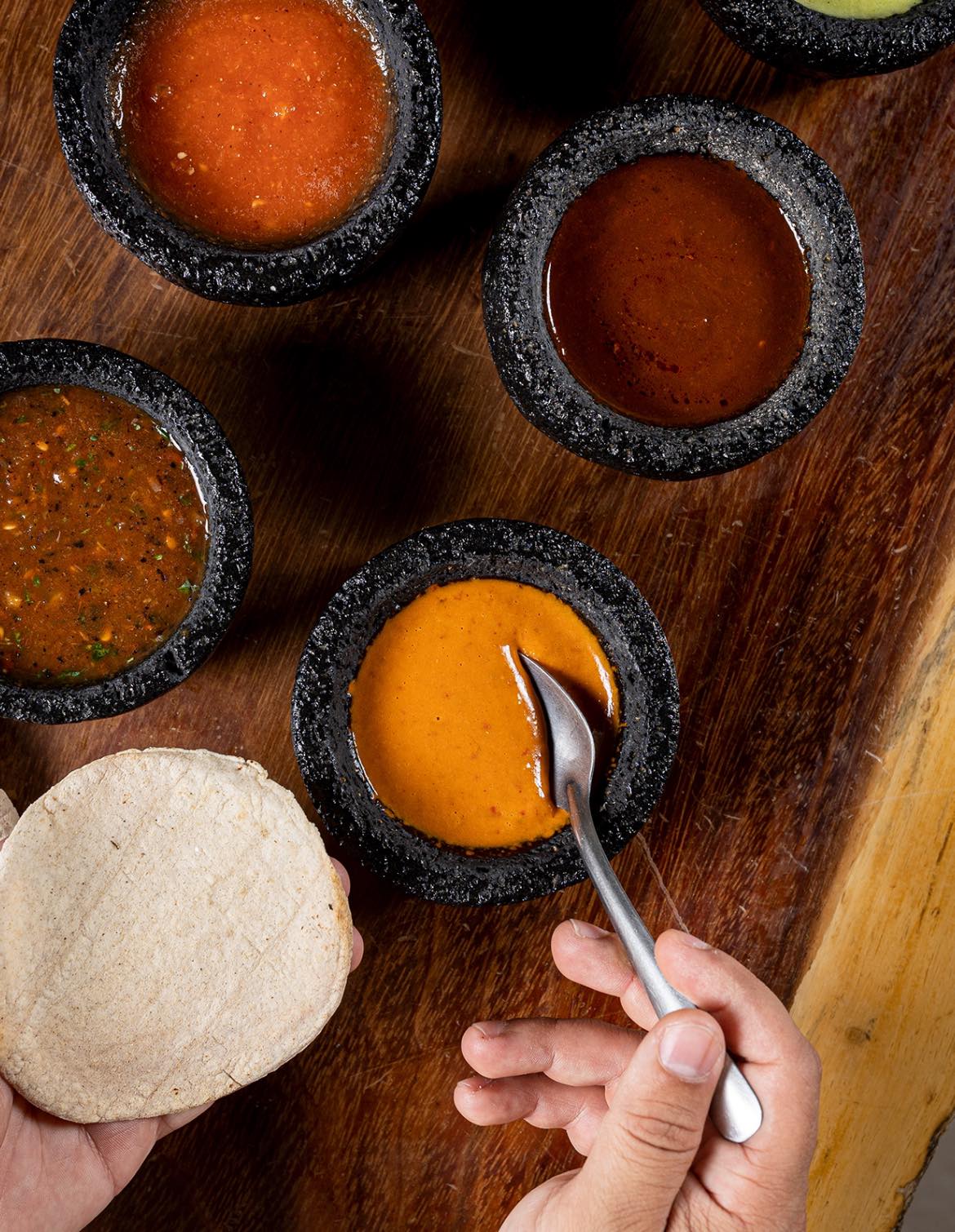 Hands holding a tortilla and spoon above a selection of colorful salsas in black stone bowls on a wooden surface.
