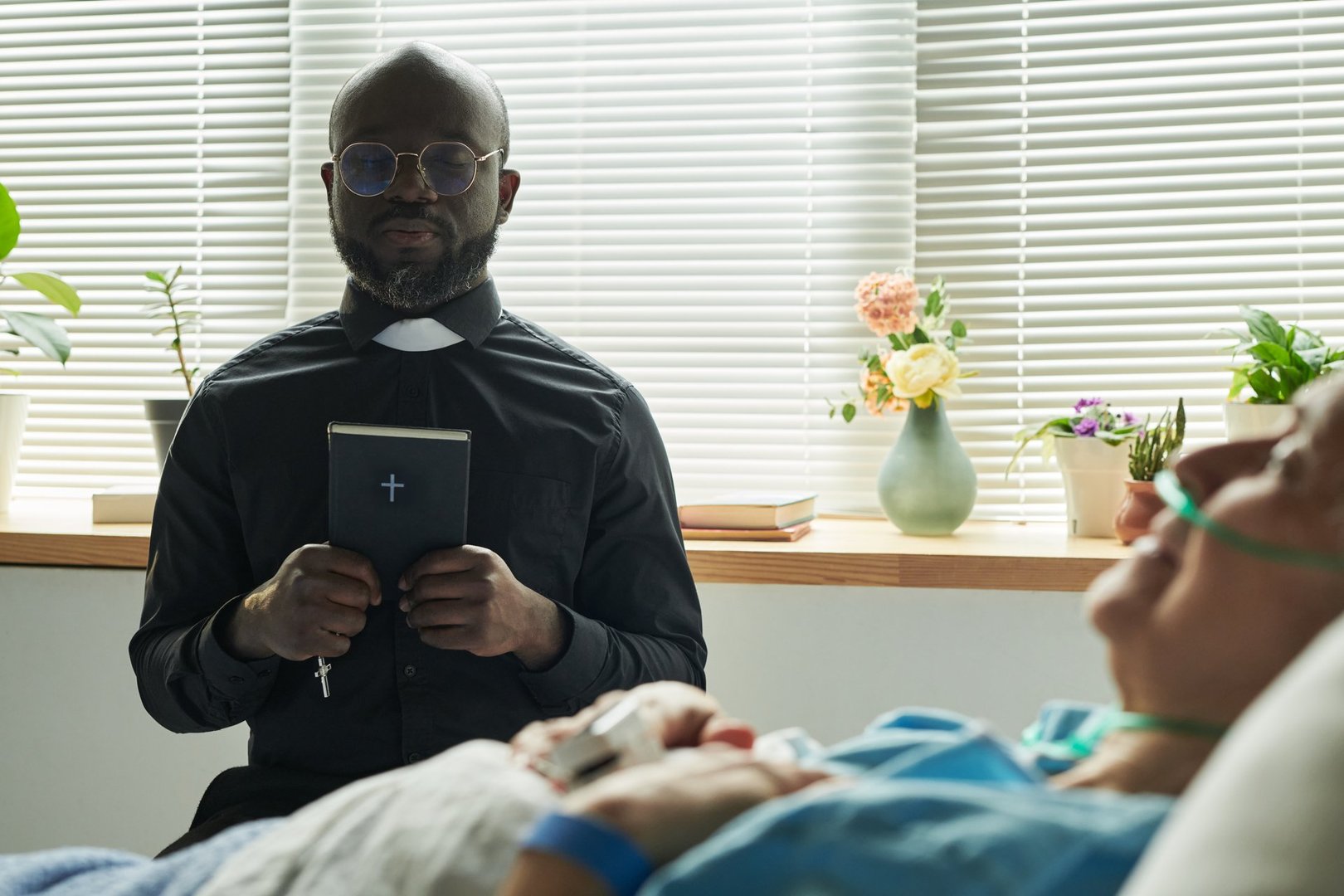 Black mature male chaplain wearing clerical collar, holding bible standing beside hospital bed, comforting senior Caucasian woman with oxygen tube during hospital visit, end of life pastoral care