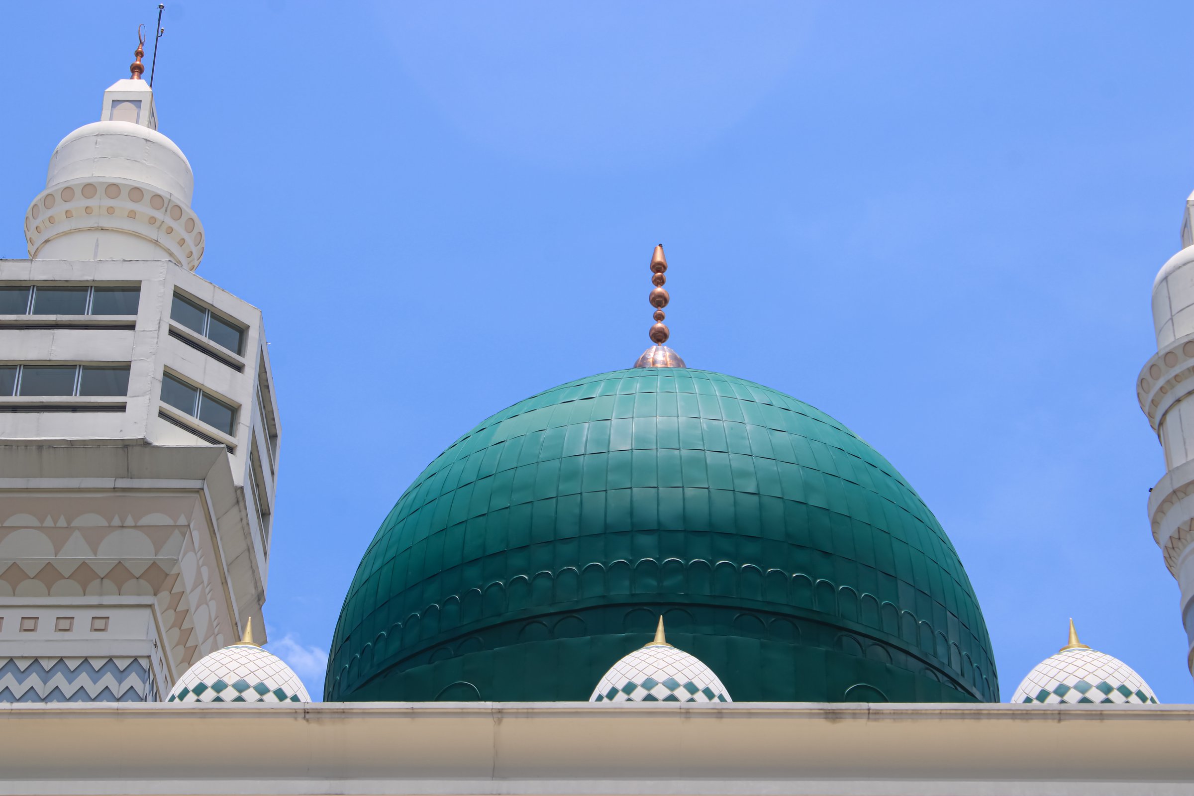 Low angle shot of a majestic mosque featuring a prominent large green dome flanked by elegant white minarets. The architectural details showcase intricate patterns and modern Islamic design against a clear, vibrant blue sky. This high-quality image captures the peaceful and spiritual essence of religious architecture. Ideal for content related to Islam, Ramadan, Eid celebrations, travel in Muslim-majority countries, and architectural photography. The clean composition and bright colors make it perfect for editorial use, banners, and cultural presentations