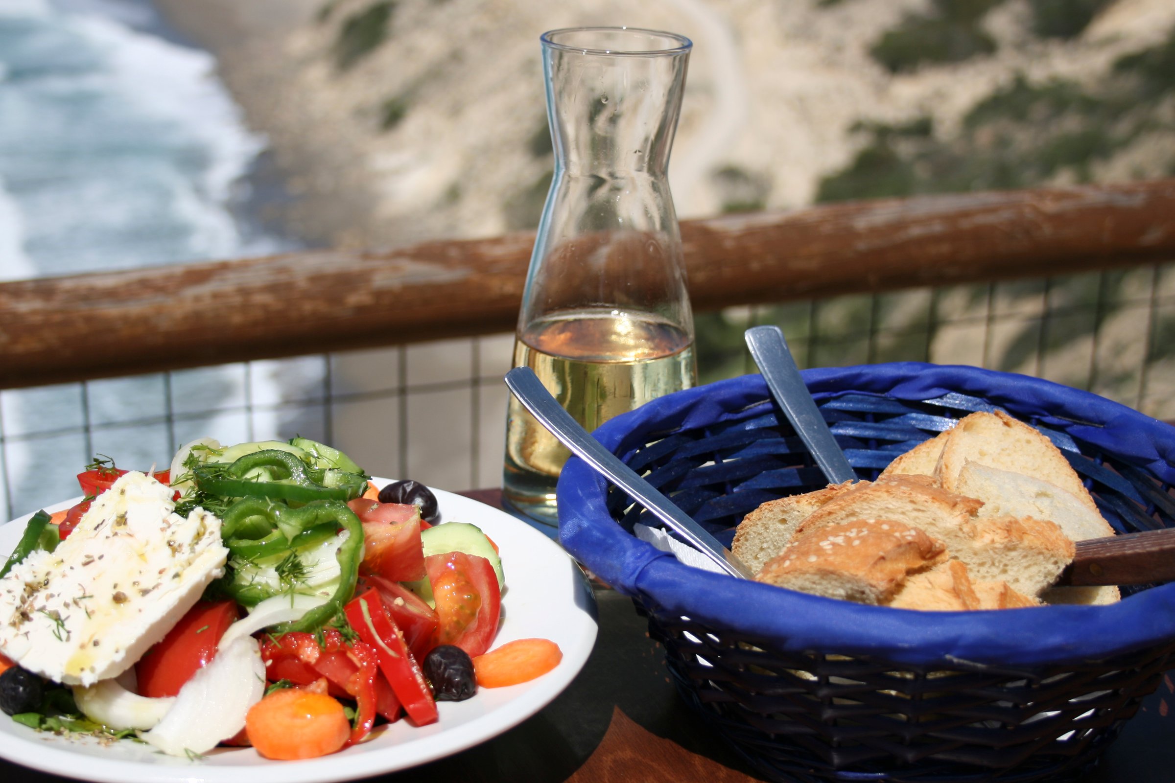 Plate of Greek salad and a basket with bread next to a carafe of white wine overlooking a coastal view.