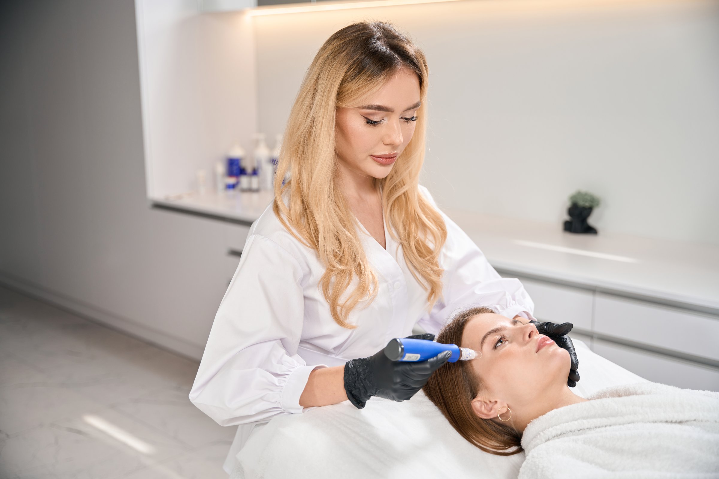 Focused female european cosmetologist doing ultrasonic cleaning of face skin with skinpen with micro needle of young caucasian woman in blurred beauty salon. Concept of face skin care and rejuvenation