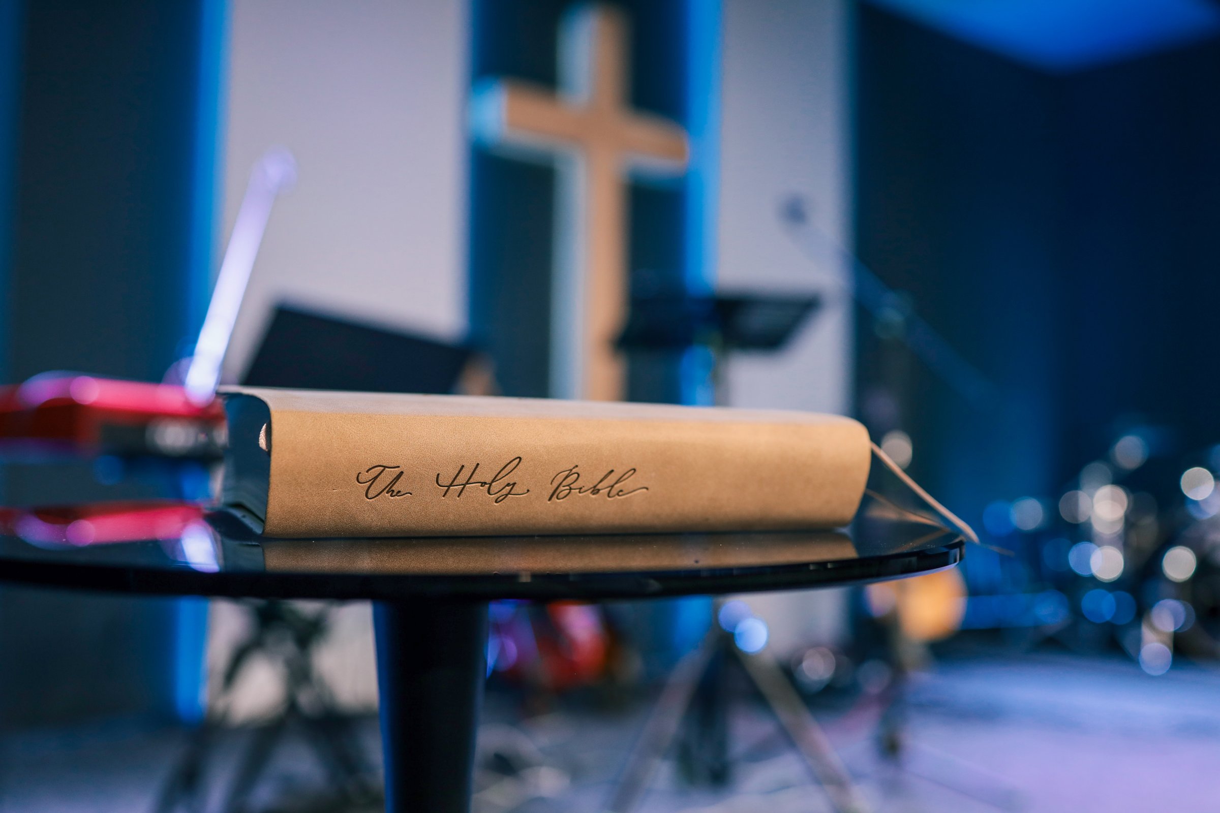 Holy Bible placed on a round black table inside a church auditorium with a wooden cross and musical instruments in the background.