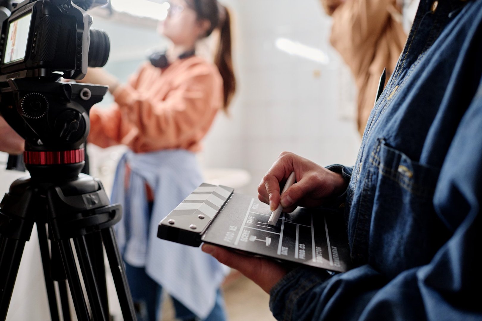 Close-up of hands holding clapperboard while filming with professional camera in indoor setting. Background showing blurred figure working with equipment