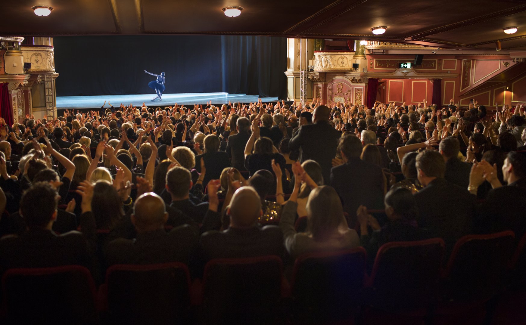 A performer takes a bow on stage in front of a standing audience in a theater with ornate interior.