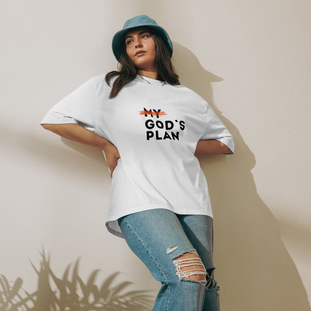 Woman wearing a white T-shirt with "My God's Plan" text, ripped jeans, and a blue hat, posing in sunlight next to leaves.