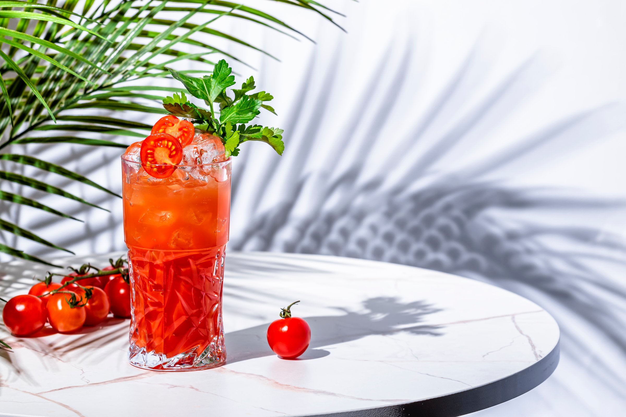 Tomato spritz alcoholic cocktail drink with gin, dry vermouth, tomato water, club soda, prosecco, cherry tomatoes and parsley. White marble table, palm leaves and shadows