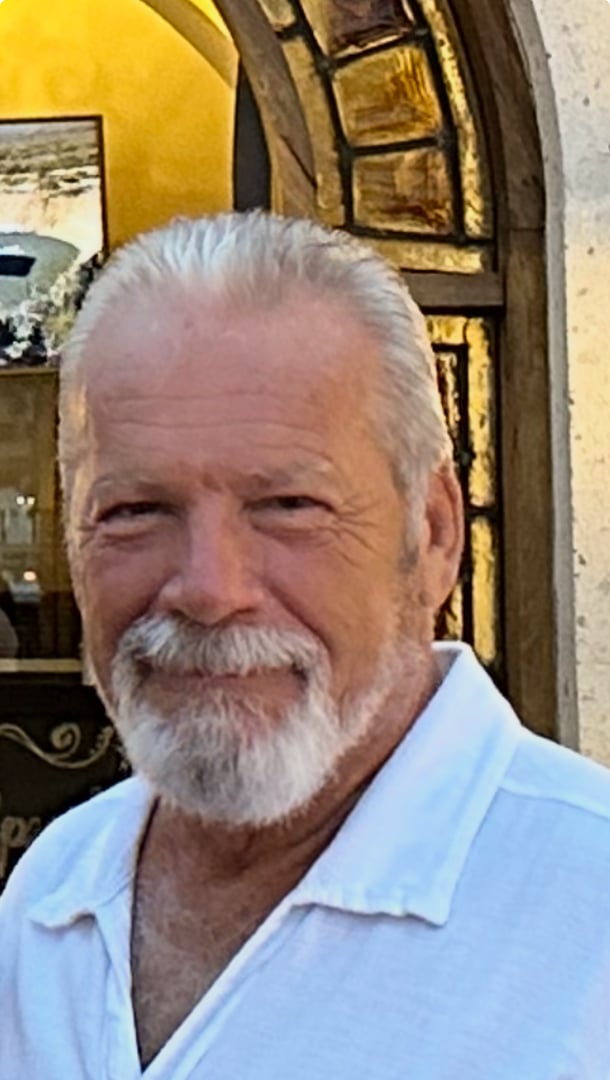 Man with white hair and beard wearing a white shirt, standing in front of a decorative archway.