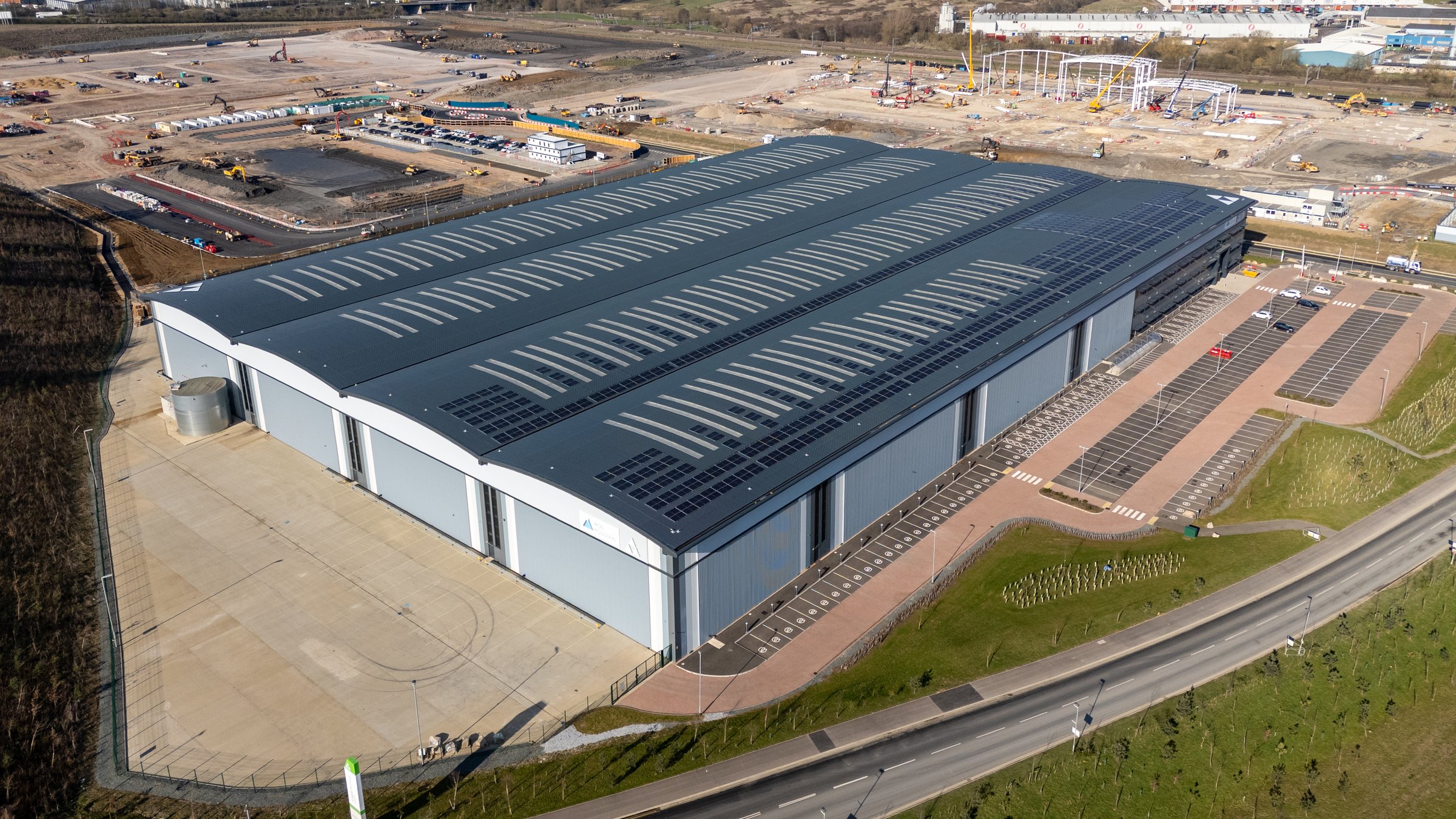 Aerial view of a large distribution center with solar panels installed on the roof, located near a highway and a construction site