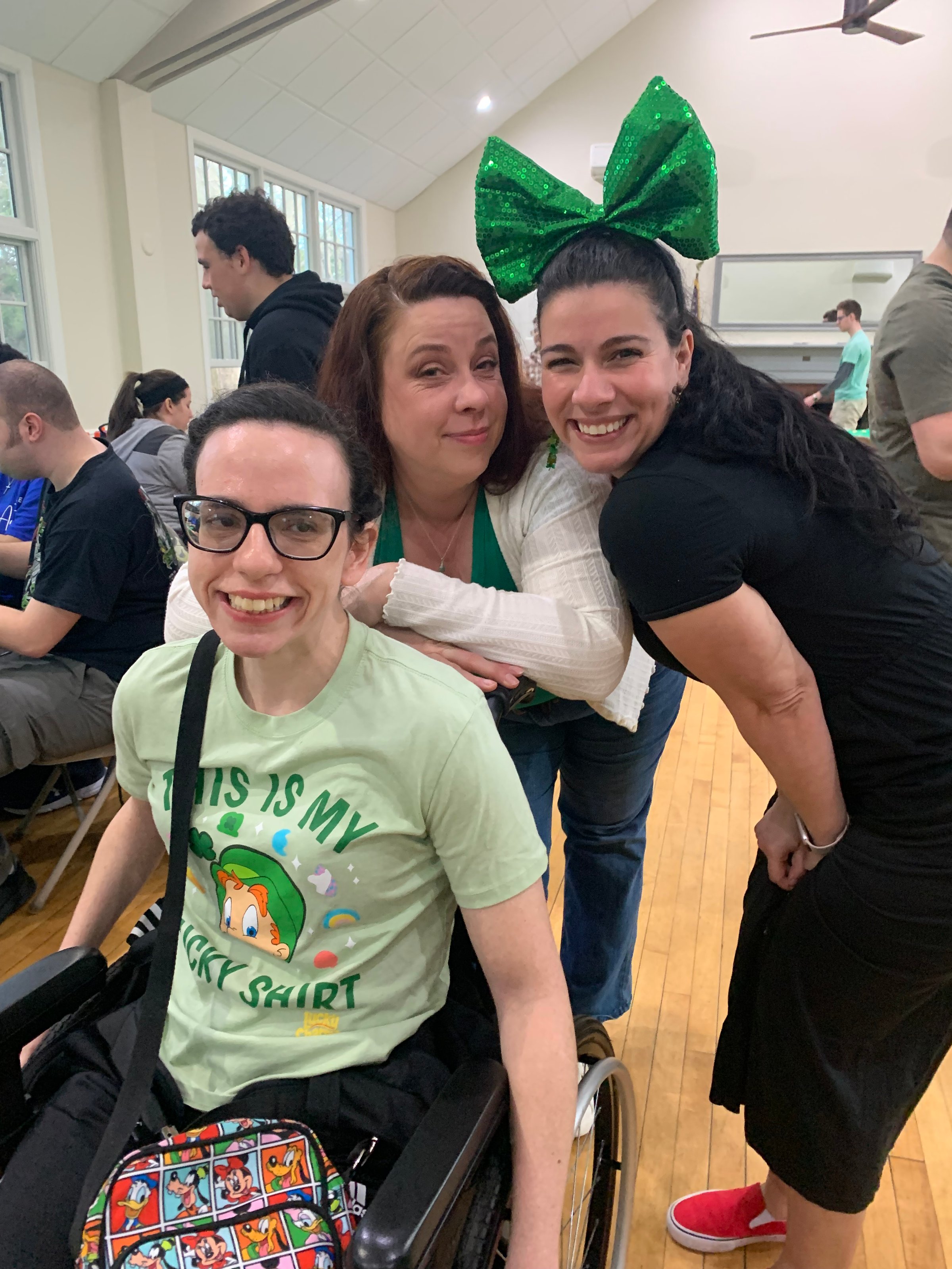 Three people smiling indoors, one in a wheelchair. Woman with large green bow. Casual setting with others in background.