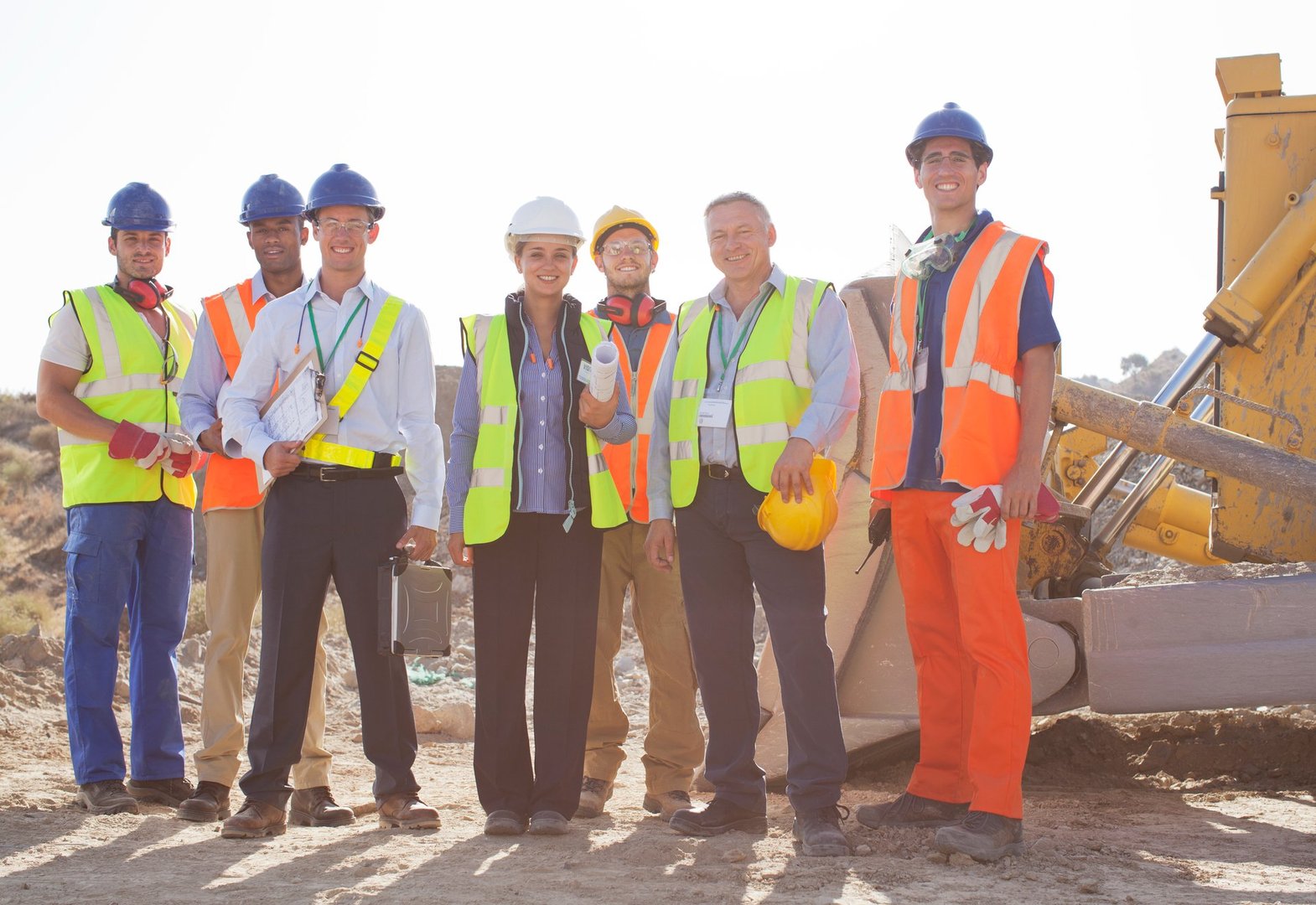 Construction team posing on site in safety gear, including hard hats and vests, standing near construction equipment.