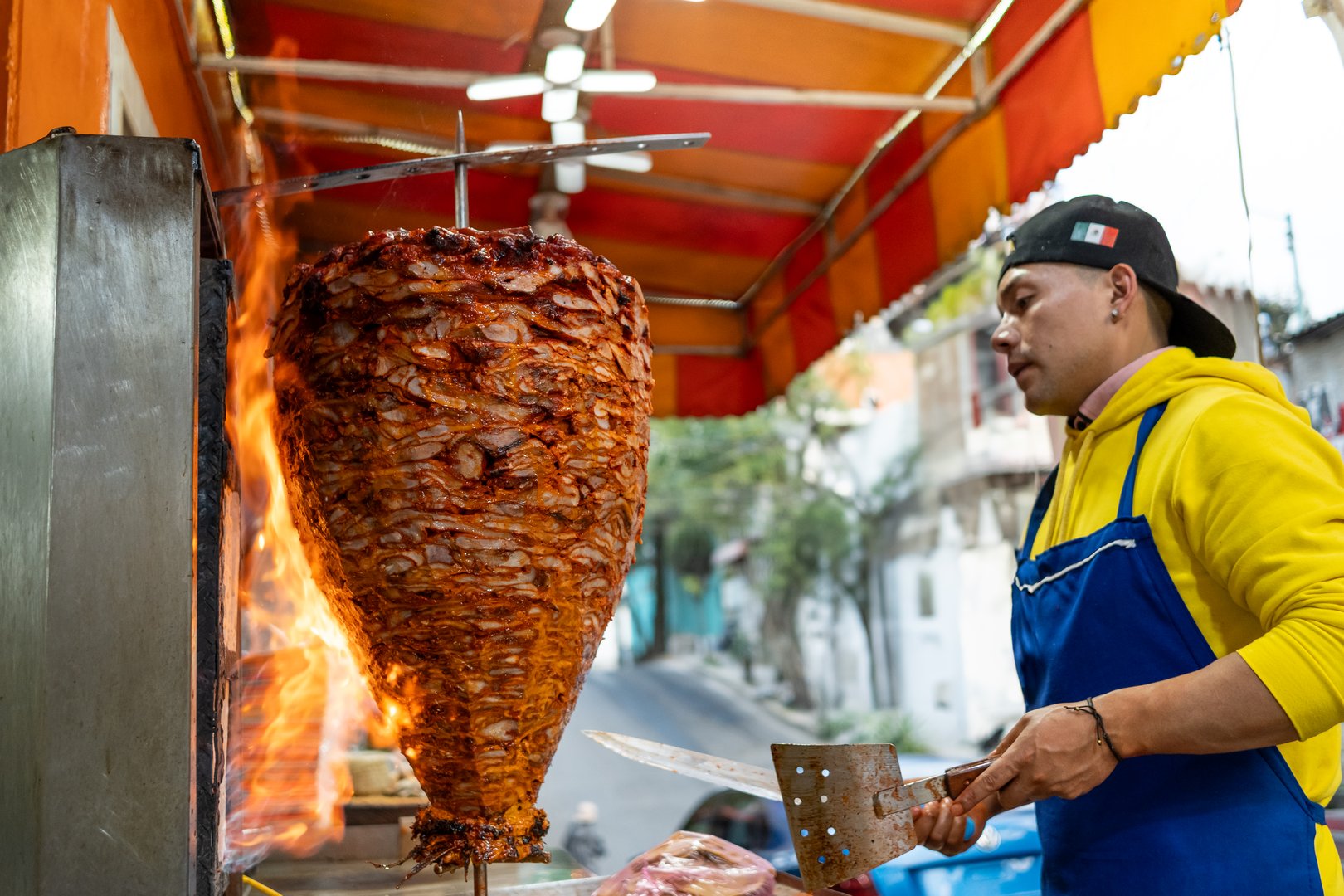 Street vendor skillfully slicing marinated pork from rotating trompo, preparing traditional tacos al pastor at vibrant street food stall