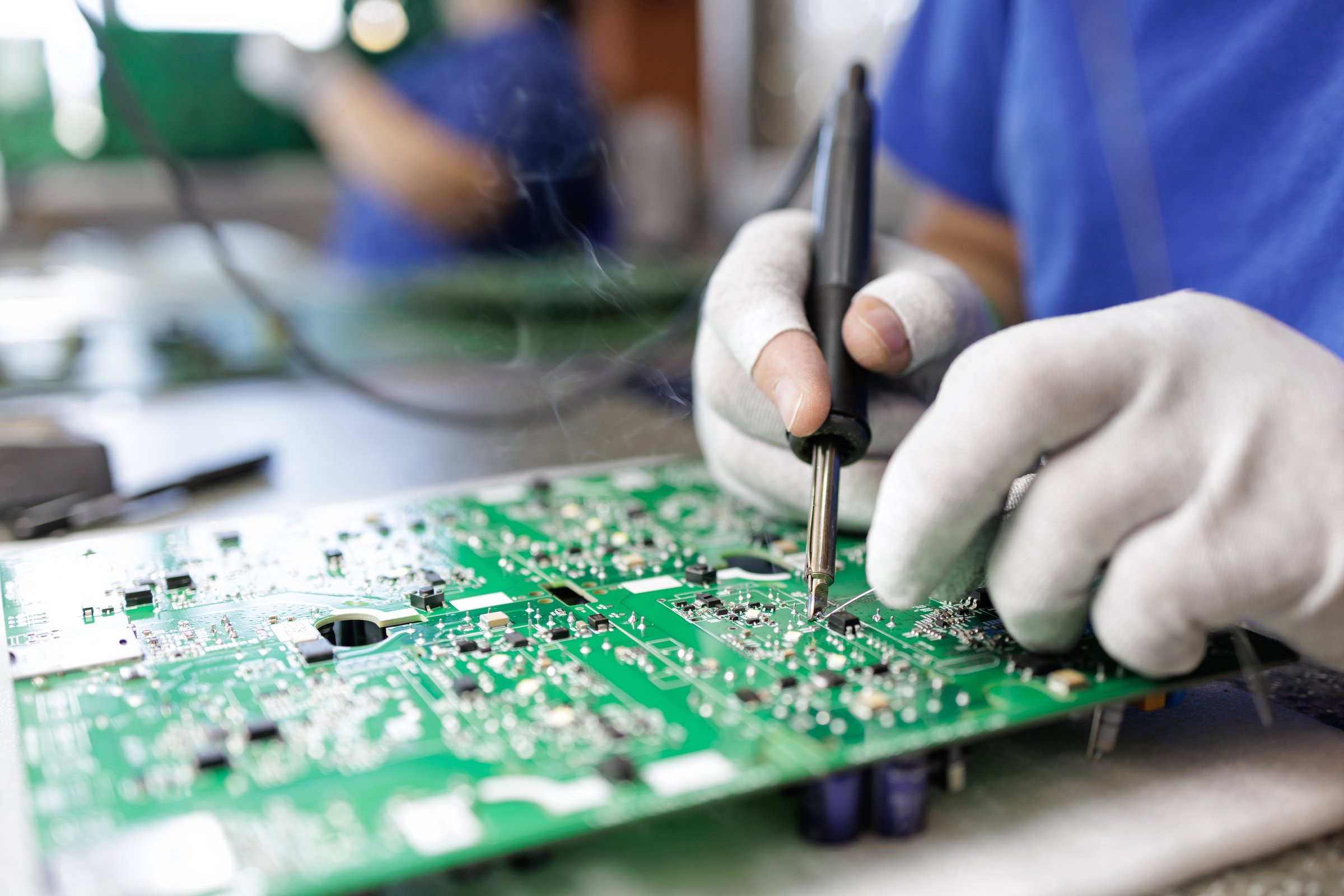 Closeup of ESD gloved worker soldering parts on a green PCB at an electronics assembly workstation in a modern factory