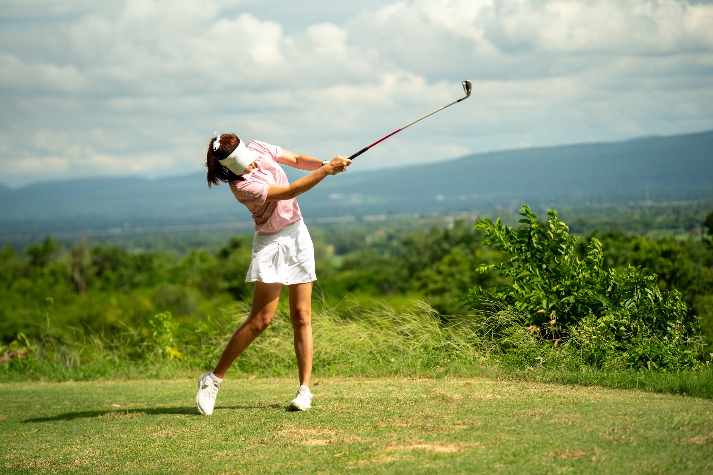 A woman is swinging a golf club on a golf course. The woman is wearing a pink shirt and white skirt