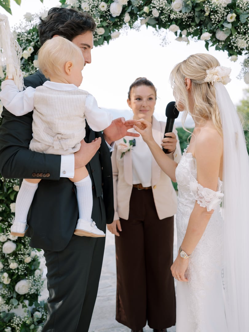 Bride and groom with a child during a wedding ceremony, standing under a floral arch, with an officiant holding a microphone.