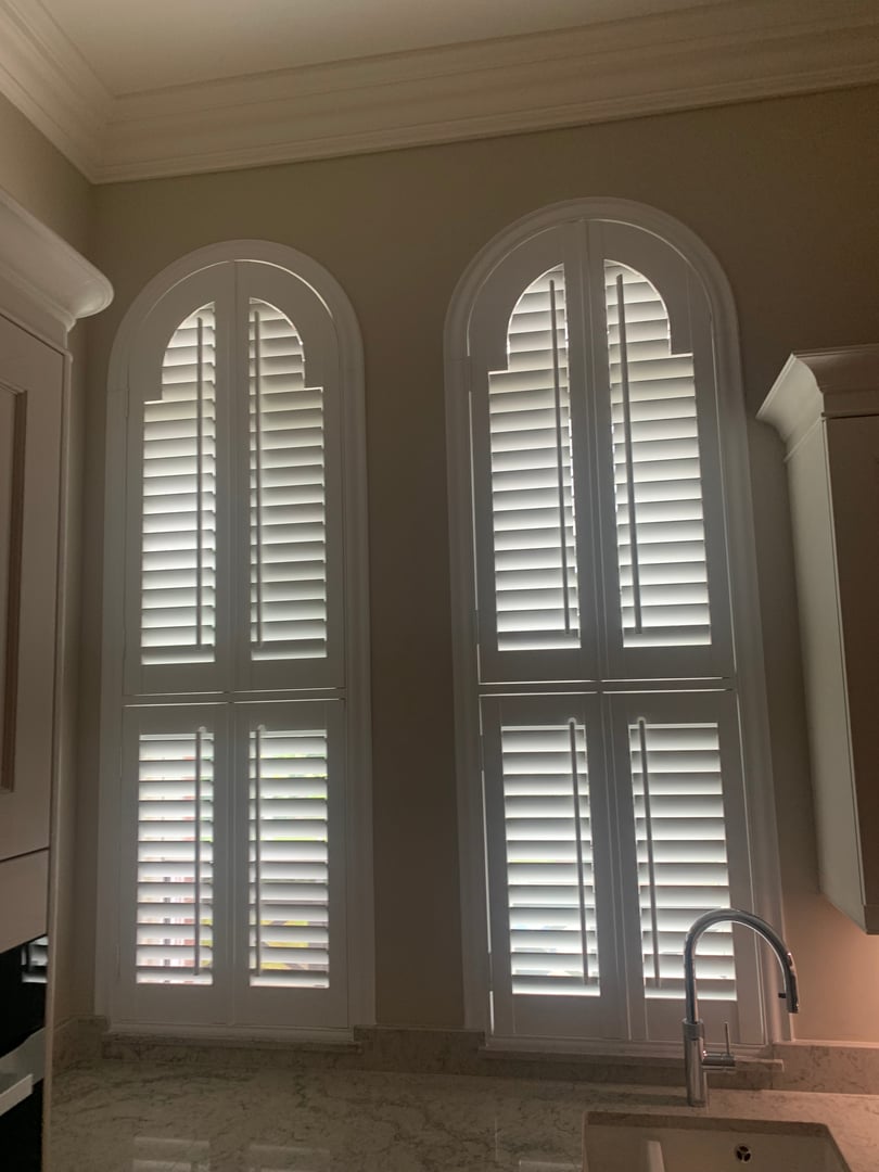 Two arched windows with white plantation shutters in a kitchen. Marble countertop and part of a sink are visible.