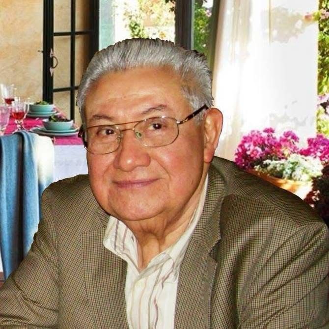 Elderly man with glasses wearing a plaid jacket, smiling indoors, with a background of potted flowers and a set dining table.