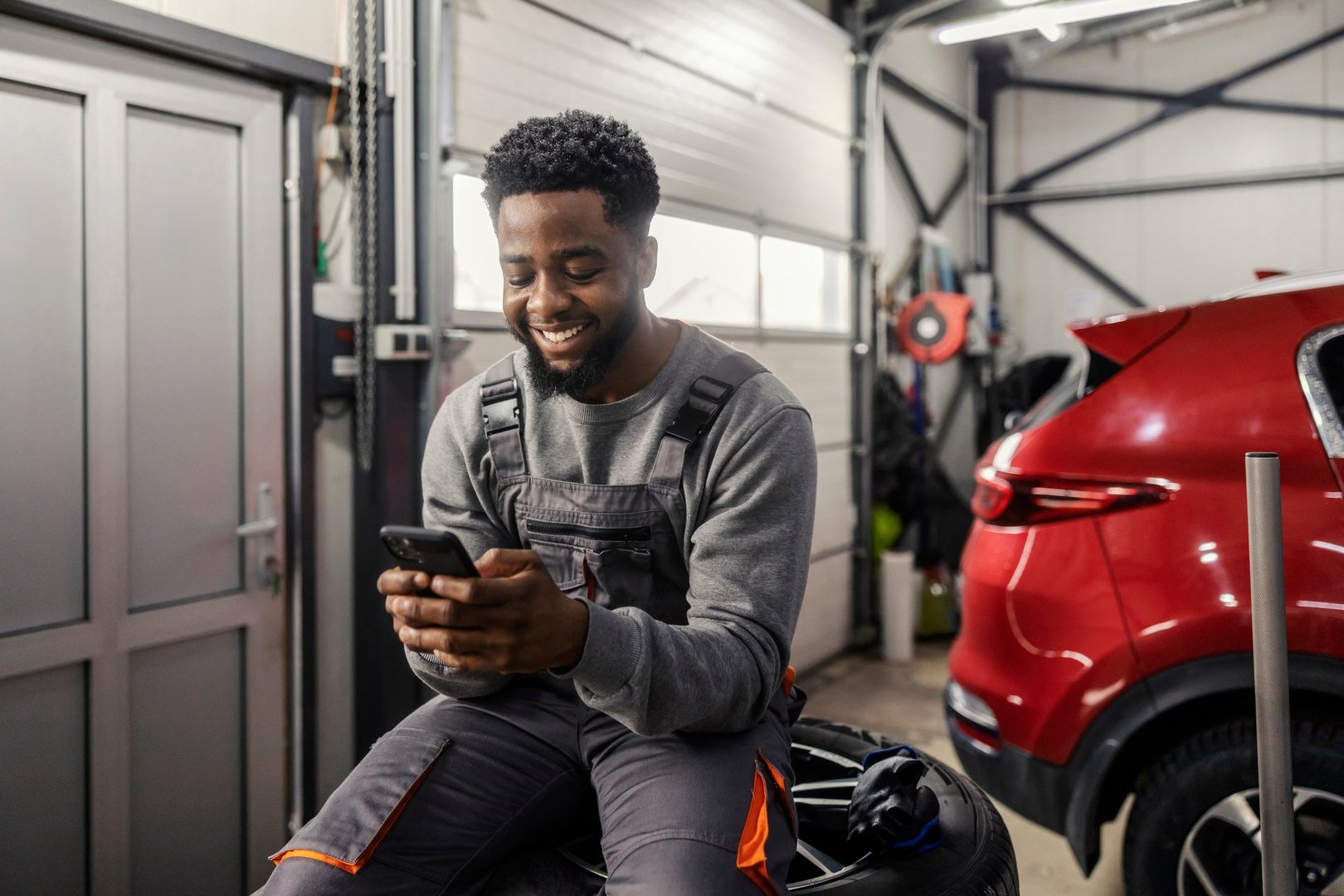Portrait of smiling interracial car service worker sitting on pile of tires and typing on his cellphone.