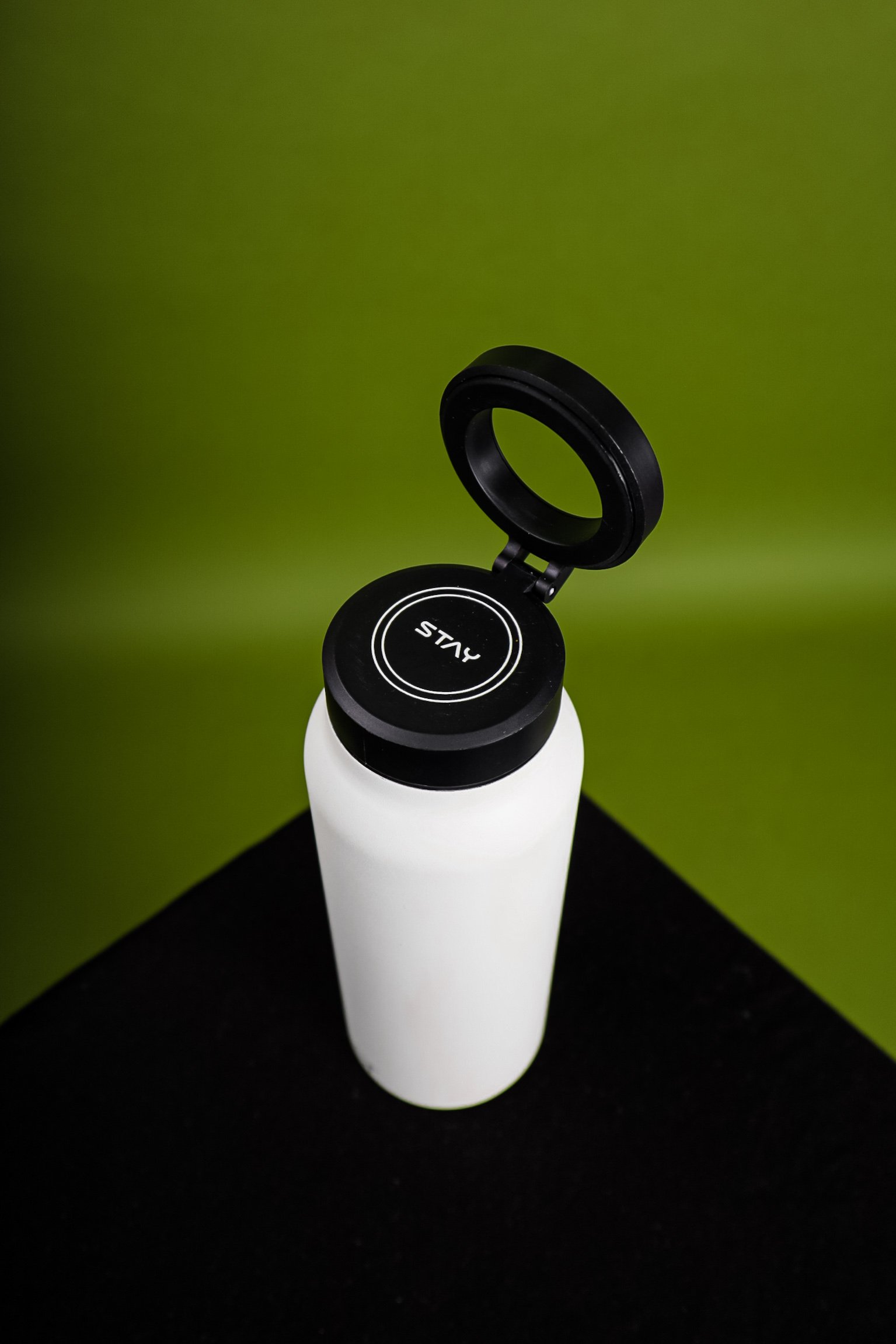 A white water bottle with a black flip-up lid on a green background. The word "STAY" is visible on the lid.