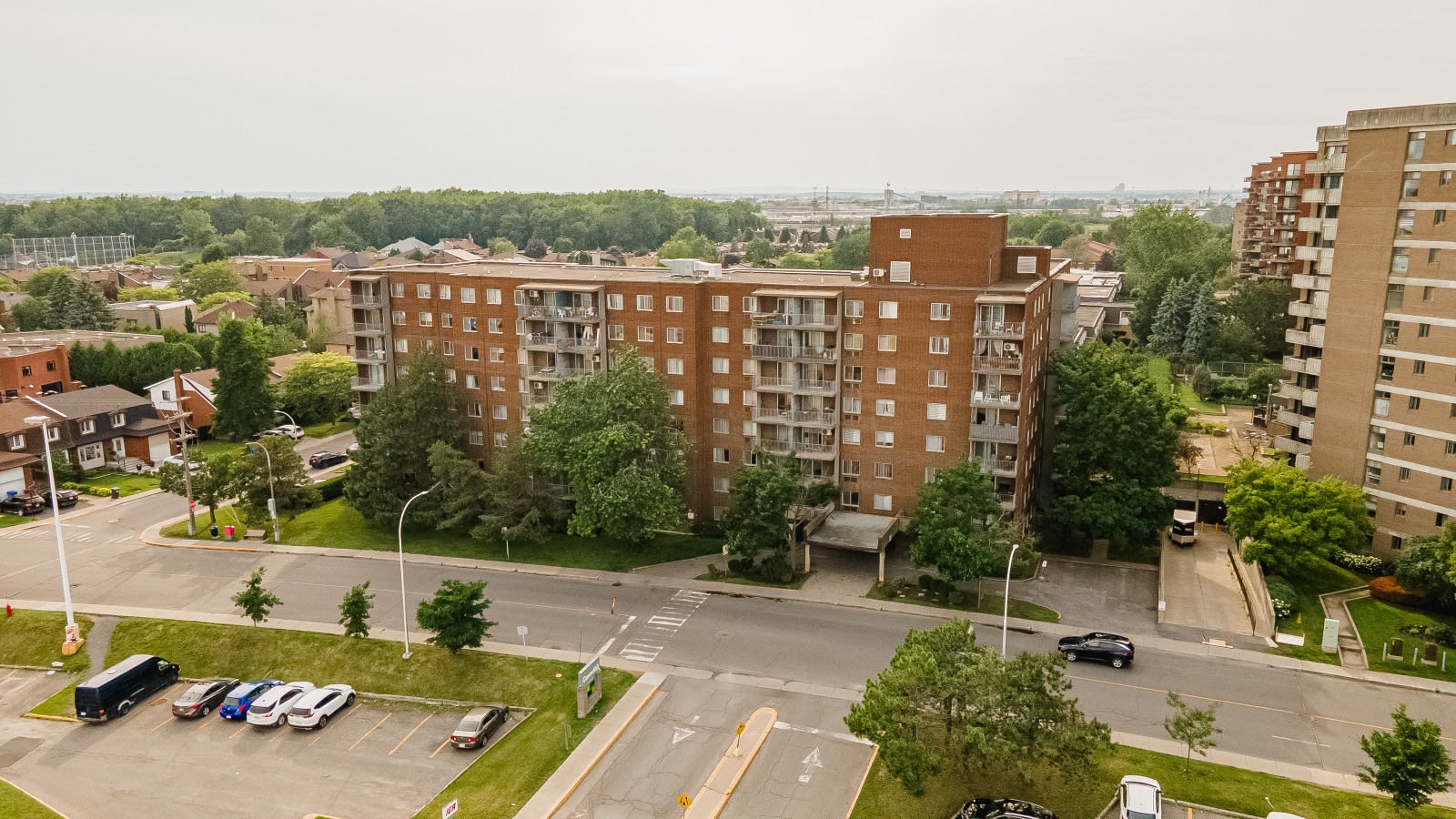 Aerial view of a mid-rise brick apartment building surrounded by trees, parking area, and roads in a residential neighborhood.