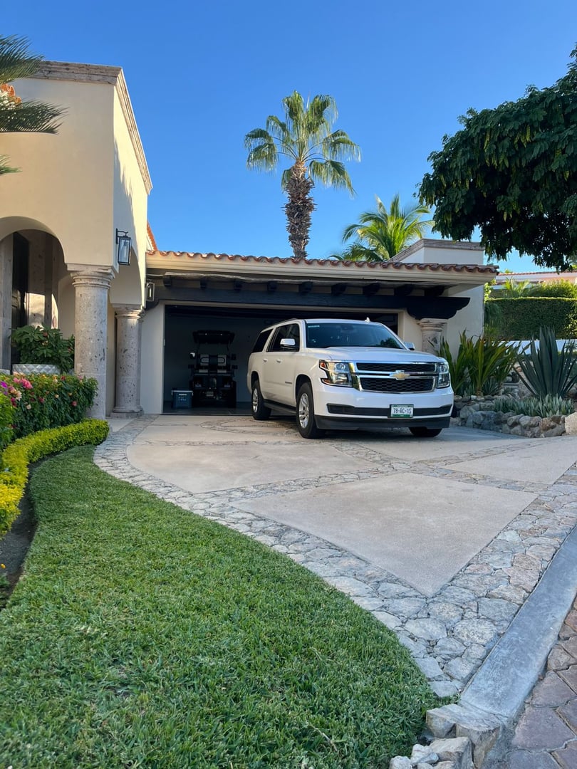 White SUV parked in a residential driveway outside a garage, surrounded by greenery and palm trees under a clear blue sky.