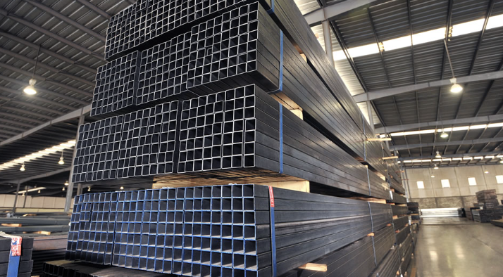 Stacks of steel rectangular tubes in a warehouse with high ceilings and overhead lighting.