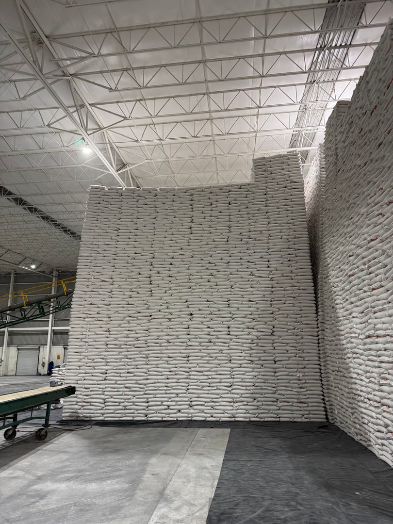 Large stacks of white bags piled high in a warehouse with a conveyor belt nearby and a metal ceiling structure above.