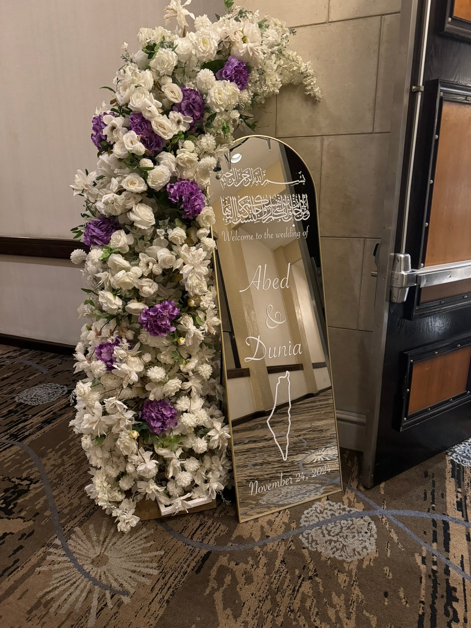 Wedding welcome sign with floral arrangement of white and purple flowers, dated November 27, 2022, alongside Arabic script.