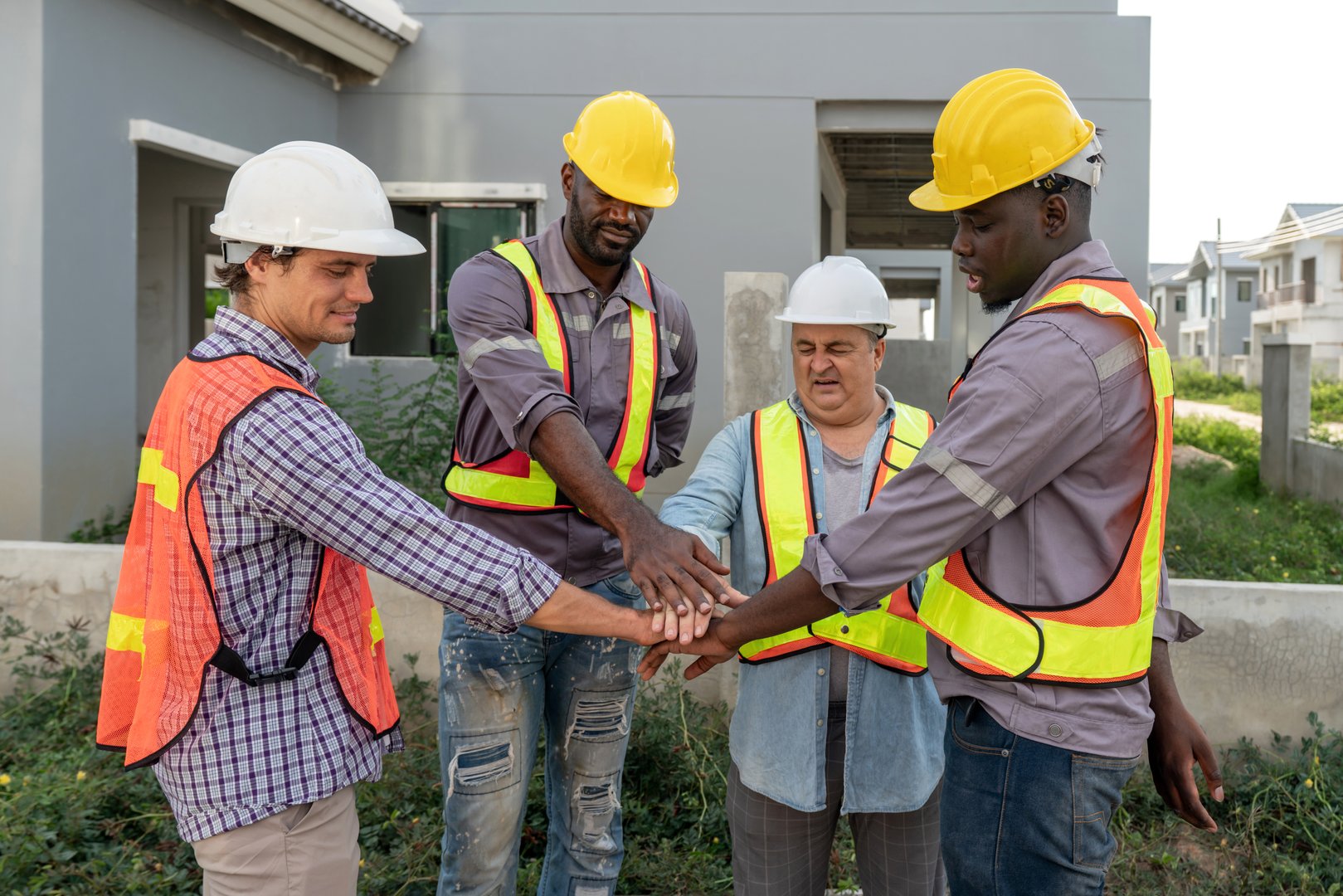 Four construction worker join hand together, stand outside a building site. They wear hardhat and safety vest.