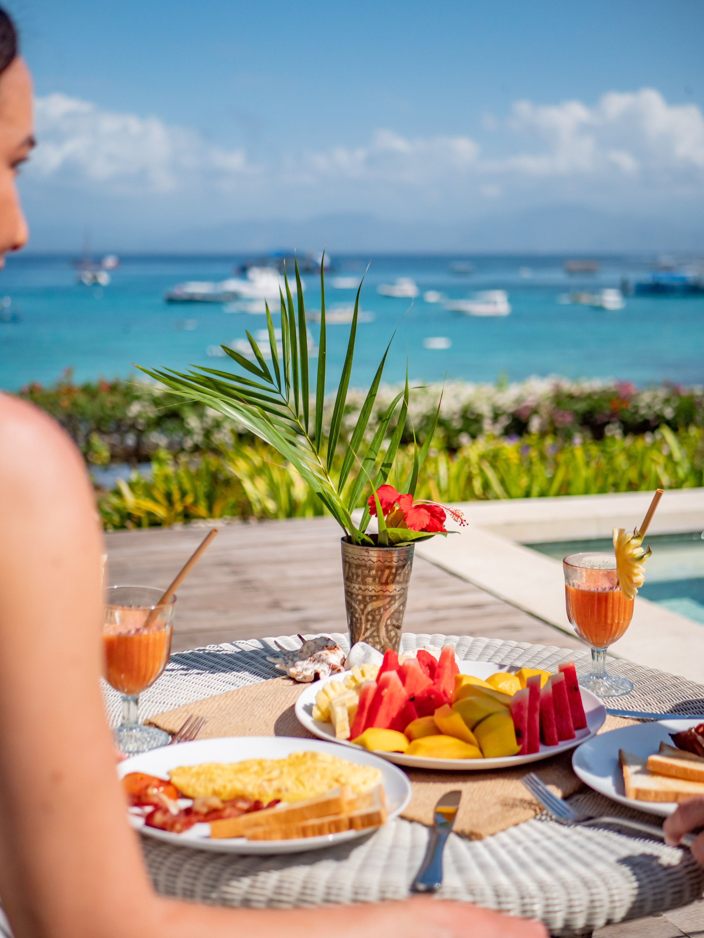 A tropical breakfast set up with fresh fruits and drinks by the seaside with boats in the background on a sunny day.
