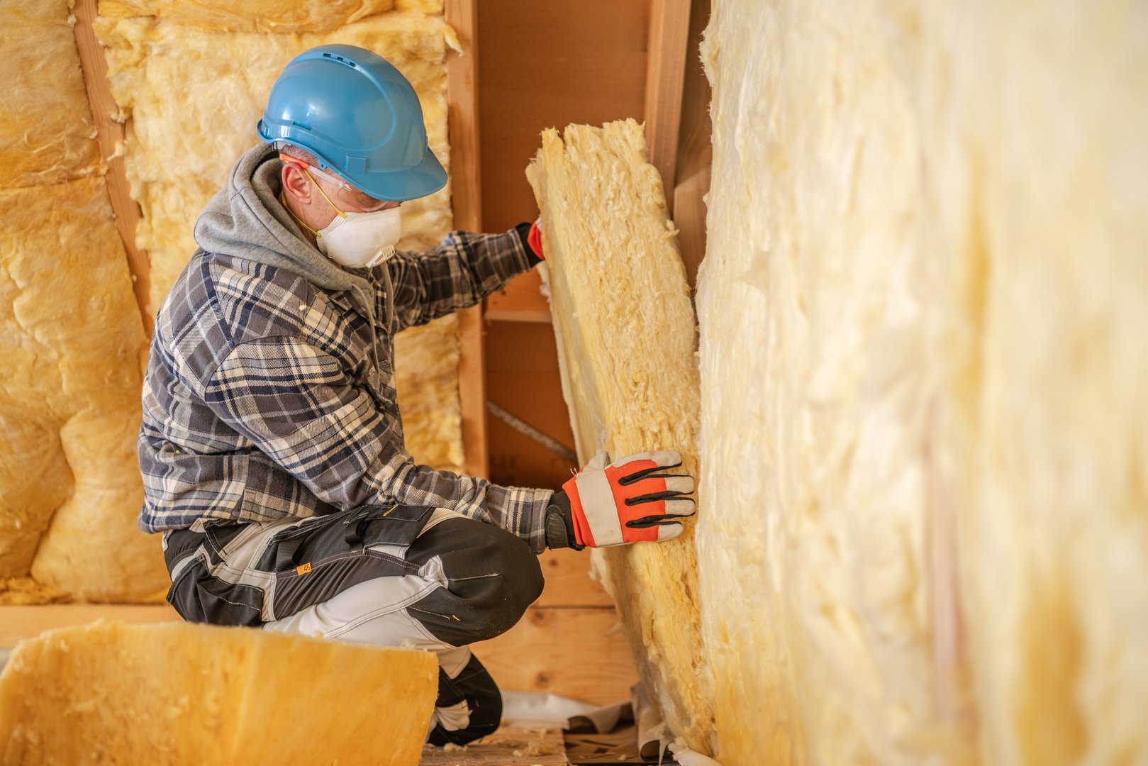 A worker wearing a blue hard hat and safety gloves is carefully placing insulation mineral wool material against a wall. The setting is an unfinished building, highlighting construction efforts