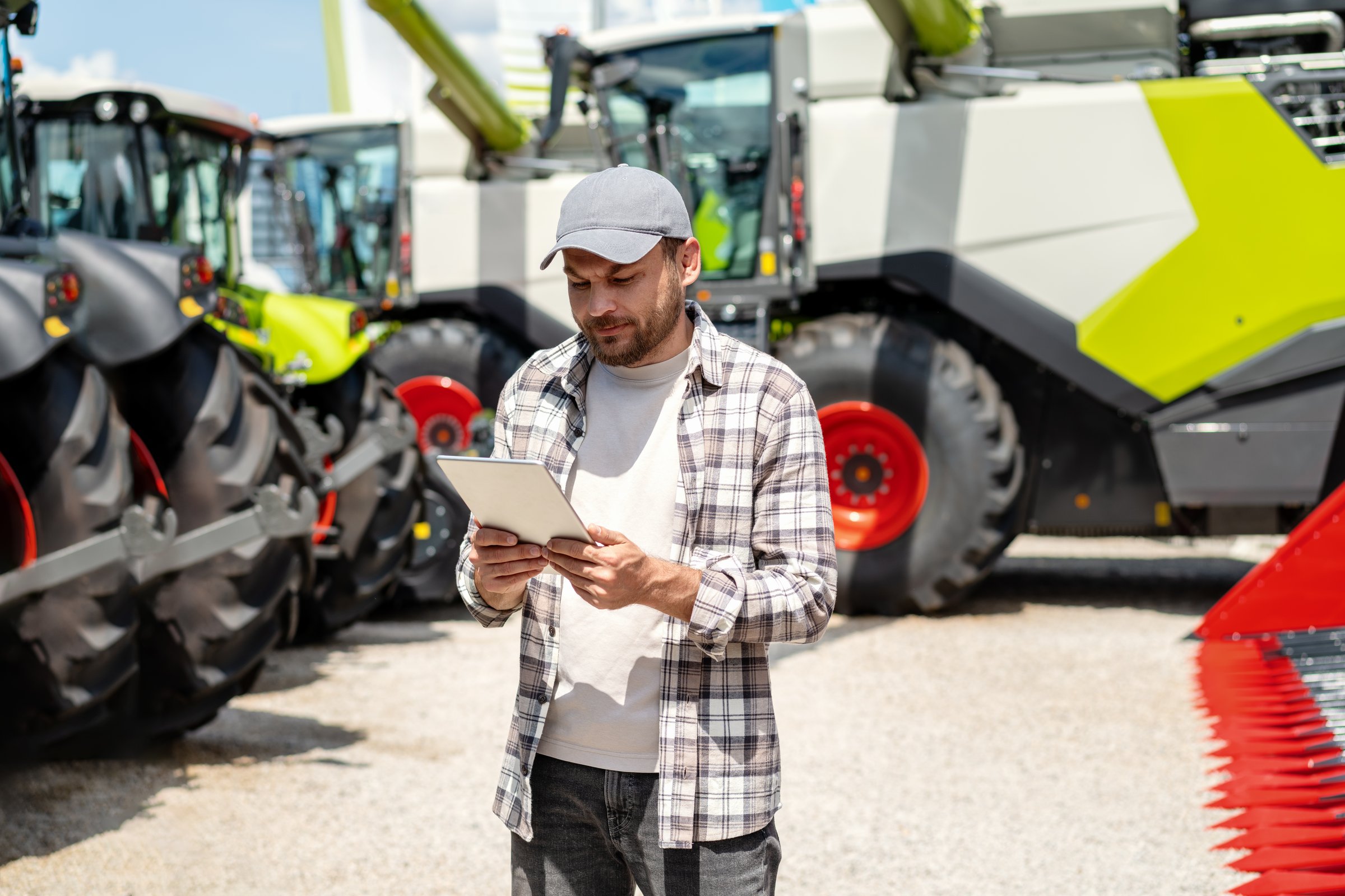 Man using digital tablet at agricultural machinery dealership. Farmer stands in front of new farm tractors and combine harvester.