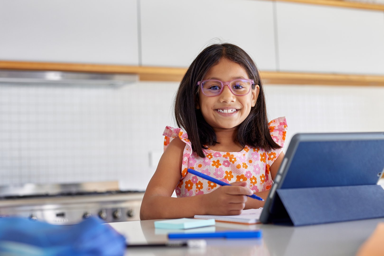 Portrait of smiling indian girl studying at home. Intelligent little girl with eyeglasses using digital tablet while writing assignment on notebook at home and looking at camera. Studious indian schoolgirl doing homework during elearning with copy space.