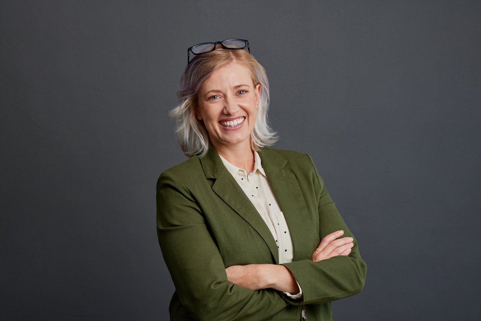 Smiling mid adult businesswoman with crossed arms looking at camera. Friendly mature woman in a casual blazer with smile and relaxed posture isolated on gray background. Confident and satisfied business woman with glasses on head.