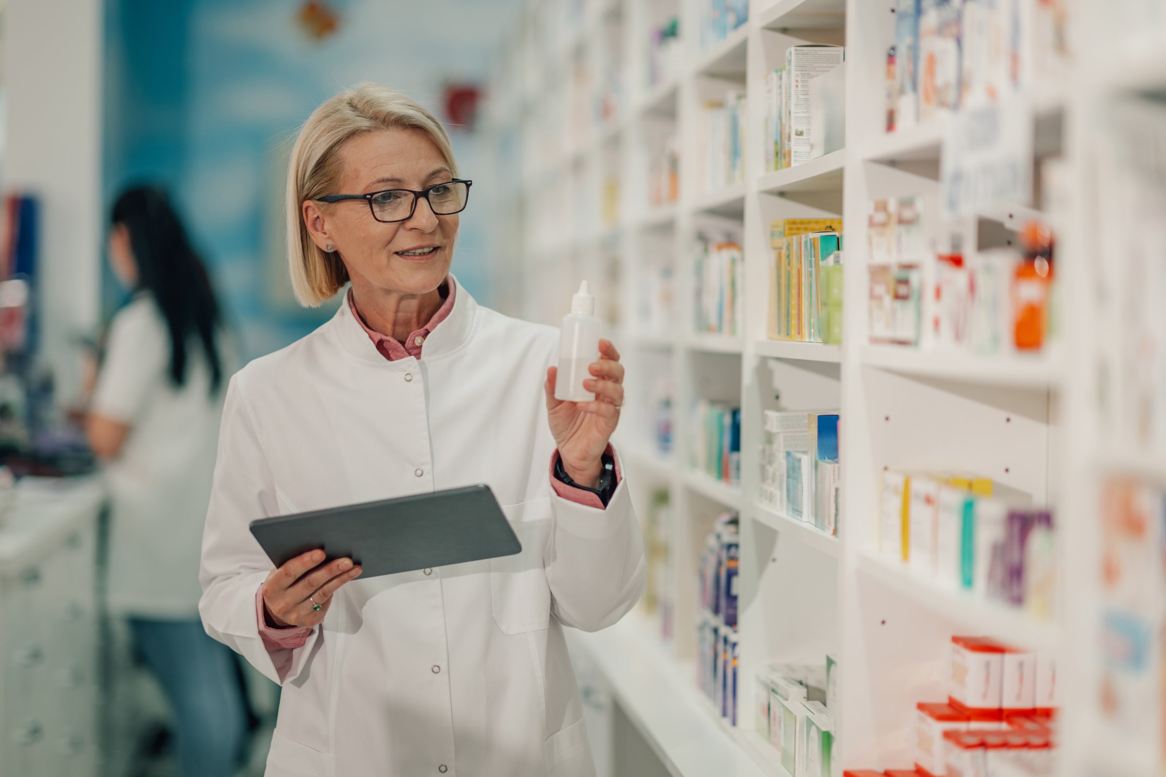 Senior pharmacist wearing uniform and holding medicine bottle and using digital tablet for checking prescription in pharmacy