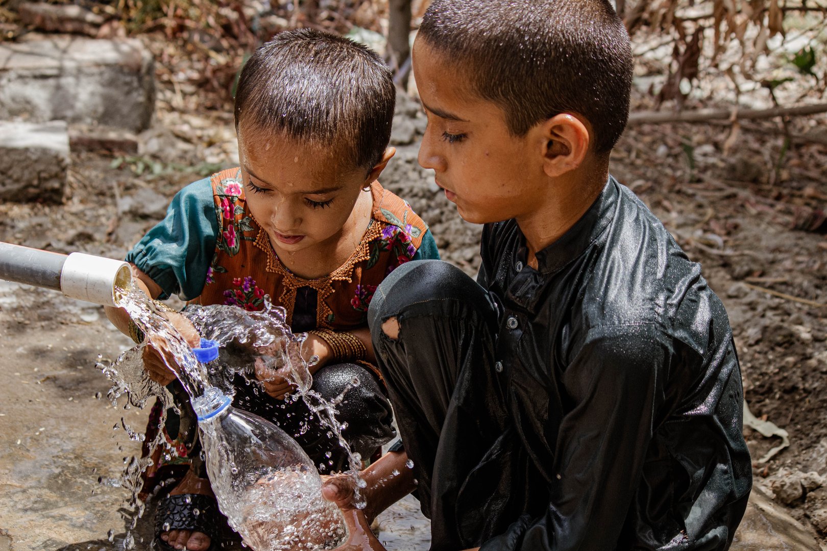 A brother and sister fill a plastic water bottle from a tap, with water splashing around them, in an outdoor setting on a hot summer day