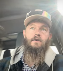 Bearded man wearing a cap and checkered shirt, sitting in a car, sunlight streaming through the window.