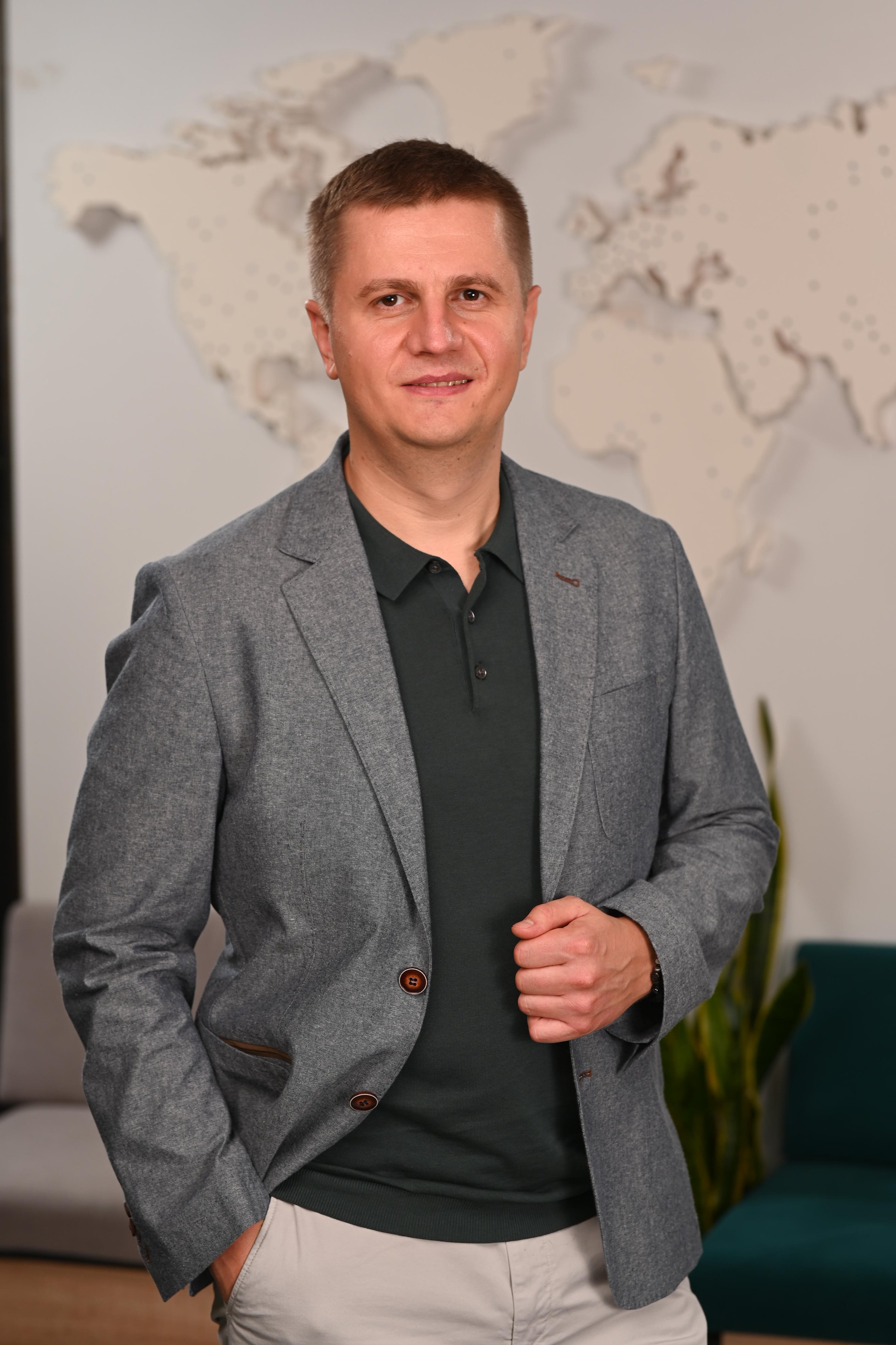 A man in a gray suit jacket and dark shirt stands confidently with one hand in his pocket, against a world map background.