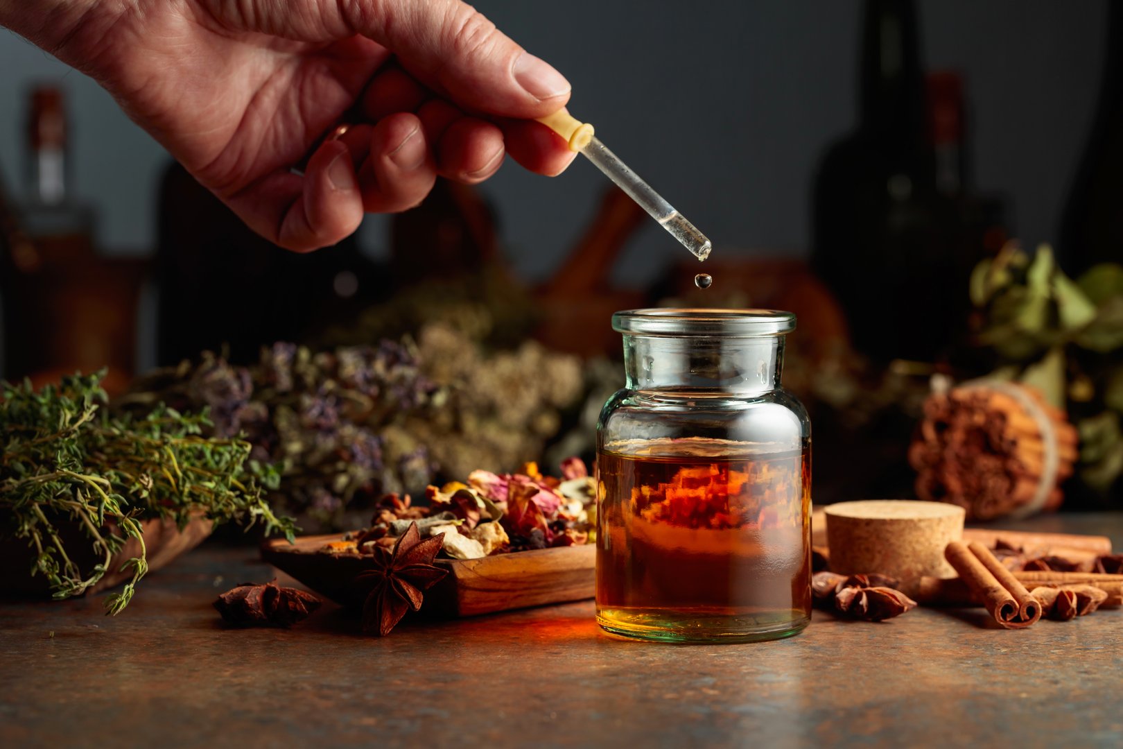Dropping essential herbal tincture into a small glass bottle. On a table dried herbs, flowers, spices, and old kitchen utensils. Concept of herbal medicine.