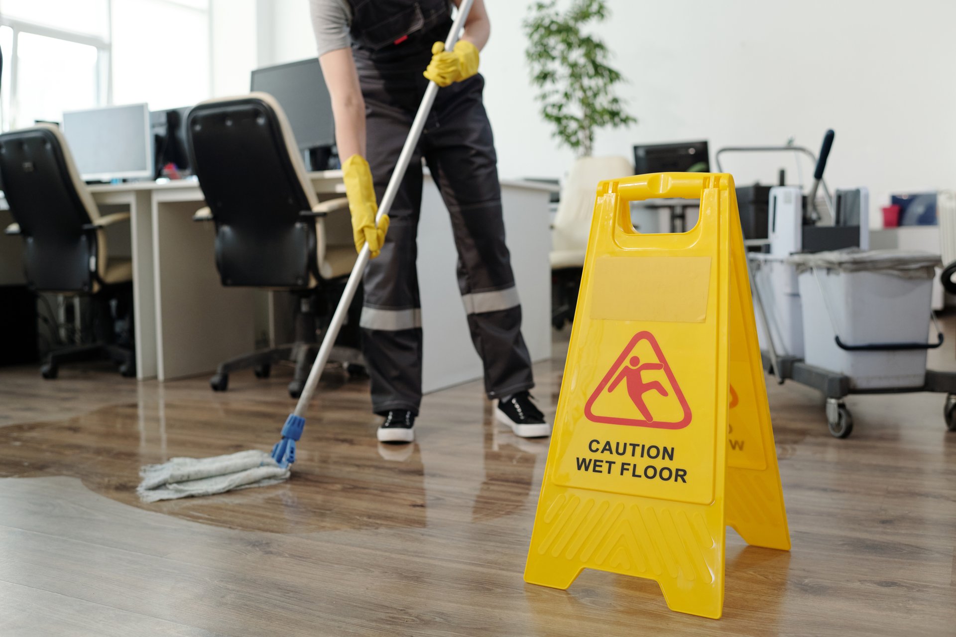 Yellow plastic signboard with caution warning about wet floor and female cleaner in black uniform working with mop in openspace office