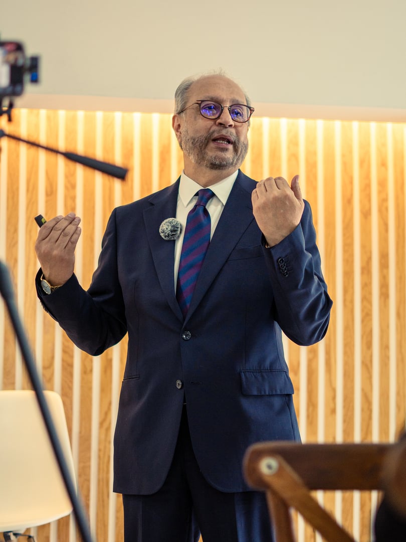 Man in a suit giving a presentation, standing in front of a wood-paneled wall, gesturing with both hands.