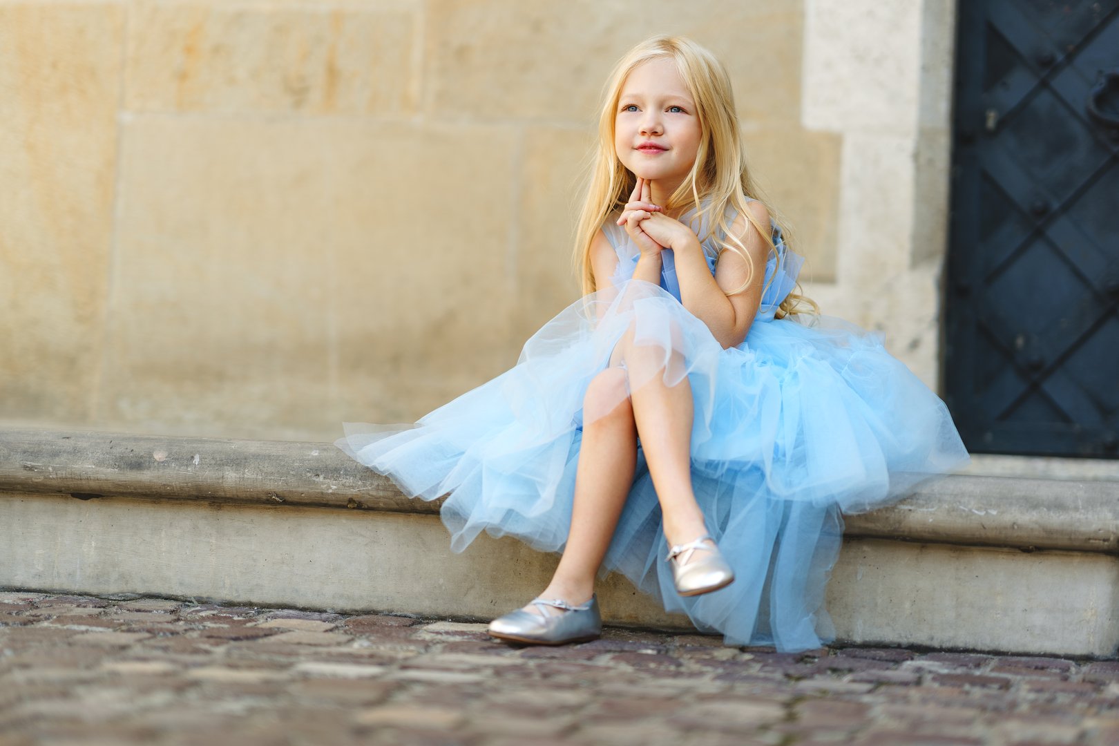A young blonde girl in a light blue dress with a bow, posing outside near a stone wall.