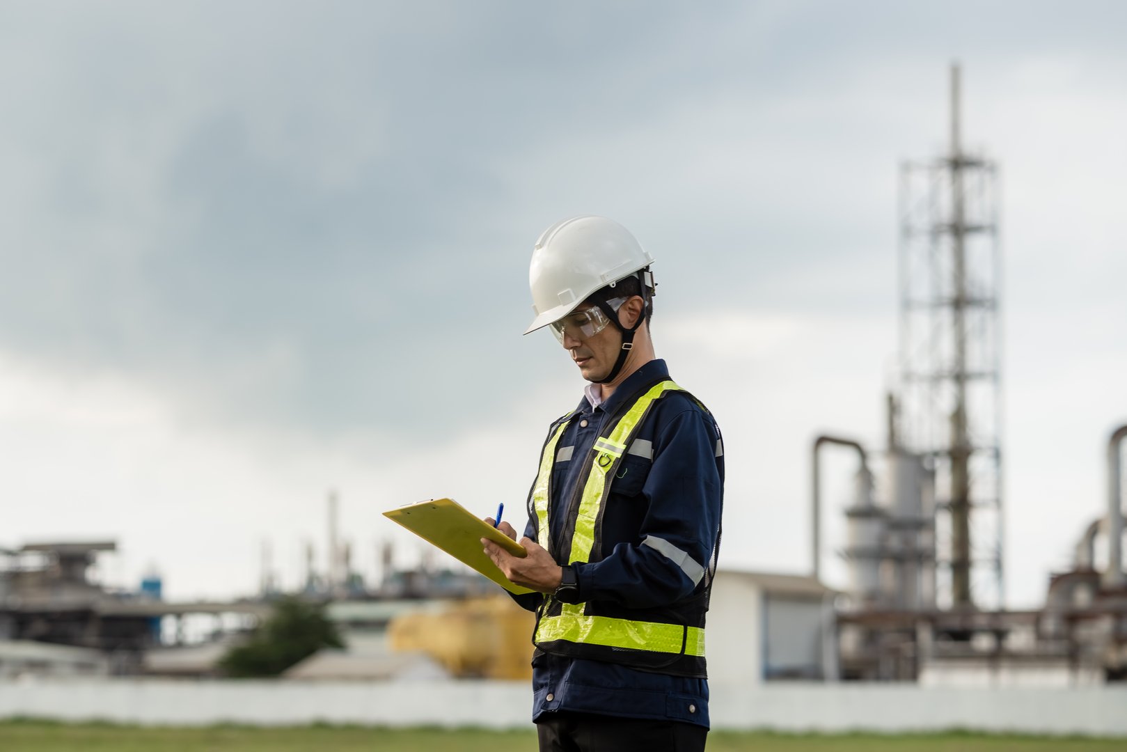 A man in a yellow vest is writing on a clipboard. He is wearing a hard hat. The clipboard is on the ground