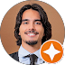 Smiling person with long hair and beard wearing a suit and tie, with a circular orange icon overlay at the bottom right.