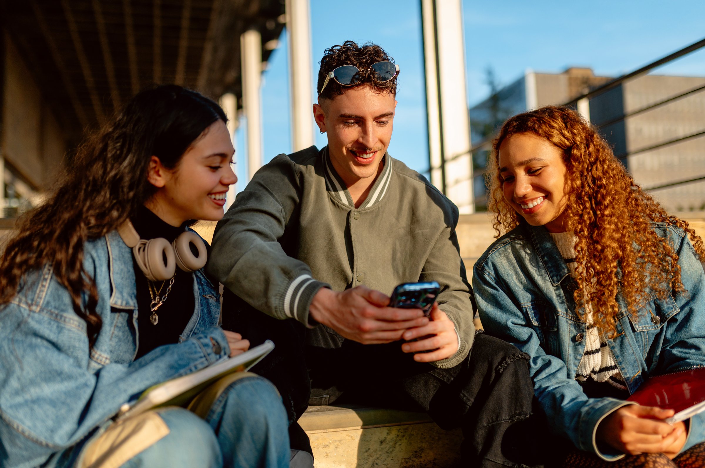 Three cheerful university students are sitting on steps, sharing a smartphone and enjoying their time together