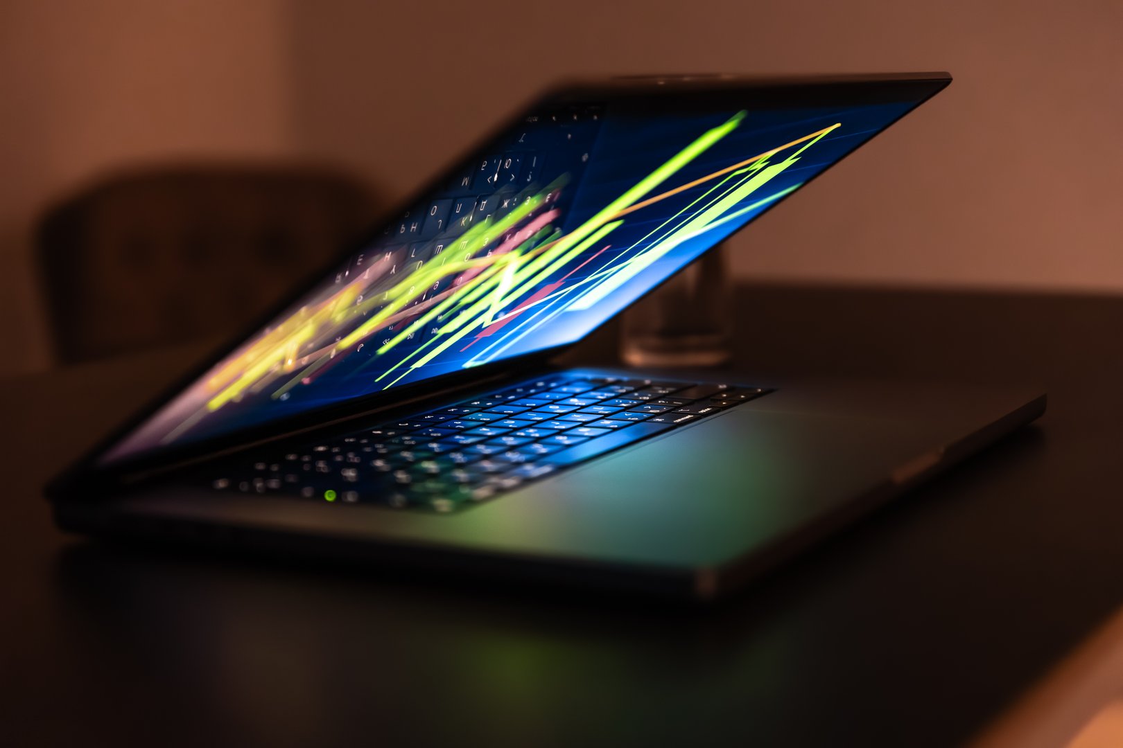 Half closed laptop on a dark desk glows with neon chart lines, the keyboard reflects cool green and blue. A scene of late analytics and remote work.