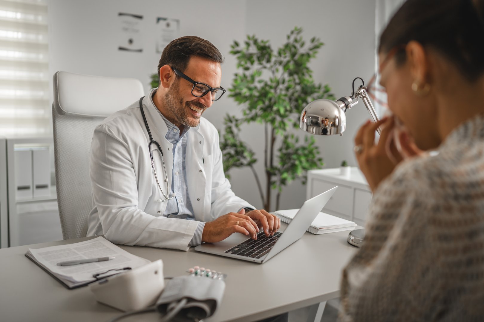Smiling doctor wearing a white coat and stethoscope laughing cheerfully while typing on a laptop during a positive healthcare consultation