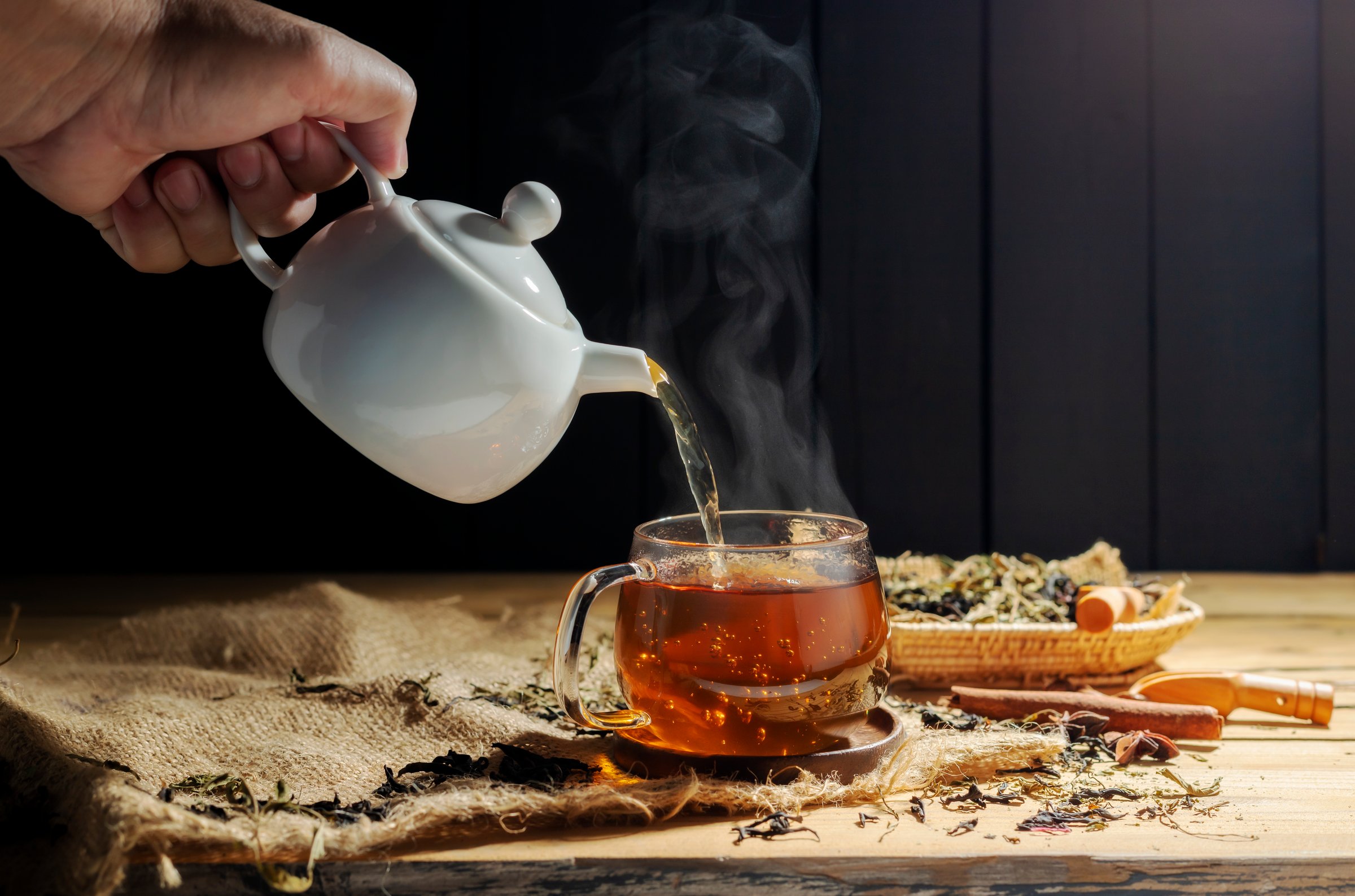 Pour hot tea into cups, herbal tea glasses are arranged on a rustic wooden table.