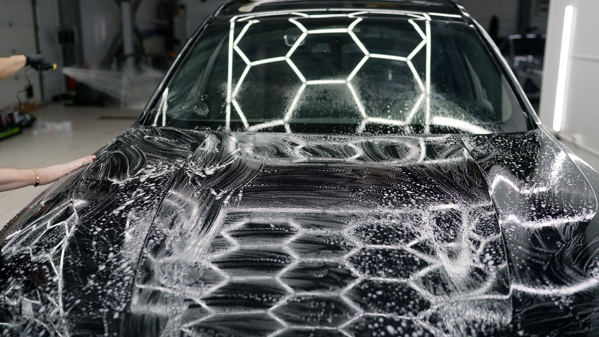 Professional car detailer applying soap and spreading foam across black car hood, meticulously preparing vehicle surface for thorough cleaning process